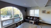 This single-floor space makes a memorable first impression with its distinctive arched window — a standout architectural feature that floods the living area with natural light and frames views of the surrounding mature trees and green landscaping. The room is finished with deep charcoal-gray walls that create a warm, intimate atmosphere, complemented by a light-toned ceiling with recessed lighting for added ambiance after dark. A dark leather sofa anchors the seating area, positioned to take advantage of the multiple windows on adjacent walls, including a traditional multi-pane window that brings in additional daylight and views of the neighboring exterior. The flooring is light in tone, providing a subtle contrast to the bold wall color. A dining or workspace table is visible in the foreground, suggesting the room serves a flexible, multi-functional purpose. A baseboard heating unit runs along the base of the arched window wall, indicating year-round comfort. The overall ambiance is cozy yet characterful, with the arched window serving as the focal point that gives the space a distinctive personality and a strong connection to the natural outdoor environment.