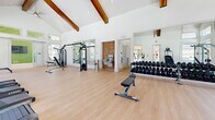 Located in Savannah, GA, this well-appointed fitness center offers a generously sized, single-floor workout facility designed to inspire and motivate. The standout architectural feature is the dramatic vaulted ceiling with rich, exposed wood beams that rise to impressive heights, creating an open and airy atmosphere throughout the space. Crisp white walls and light-toned hardwood-style flooring contribute to a clean, modern aesthetic that feels both inviting and energizing.

Natural light pours in from an extensive array of large windows and glass doors positioned along multiple walls, offering views of the surrounding greenery and outdoor areas. A living green wall accent adds a refreshing natural element to the interior, complementing the lush outdoor scenery visible from within.

The cardio zone features a row of treadmills, elliptical machines, and other aerobic equipment, each positioned to take advantage of the outdoor views. The strength training area is equally well-equipped, with a multi-station cable machine, a Smith machine with bench, a full dumbbell rack, and additional free weight stations. Wall-mounted mirrors expand the sense of space and allow for proper form checks during workouts. A wall-mounted flat-screen television and a drinking fountain add convenience. Barn-style sliding glass doors provide stylish interior separation, rounding out this thoughtfully designed fitness facility.