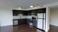 This single-floor space showcases a well-appointed kitchen designed with a contemporary aesthetic. Dark espresso-toned cabinetry lines the walls in an L-shaped configuration, providing generous storage through both upper and lower cabinets fitted with sleek bar-pull hardware. The cabinetry is complemented by a light-colored subway tile backsplash, creating a striking contrast that adds visual depth to the space.

The kitchen is equipped with a full suite of stainless steel appliances, including a French door refrigerator, a freestanding electric range with a matching over-the-range microwave, and a dishwasher. A stainless steel undermount sink is positioned along the perimeter counter, accompanied by a modern faucet. The countertops feature a dark stone or granite-style finish that coordinates cohesively with the overall color palette.

Underfoot, rich dark hardwood-style flooring extends throughout, reinforcing the modern tone of the space. Flush-mount ceiling light fixtures provide even, ambient illumination. The clean lines, cohesive finishes, and functional layout combine to create a polished, move-in-ready kitchen environment.