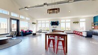 This dynamic, single-floor community amenity space is designed to foster social interaction and recreational activity in a visually stimulating environment. The open-concept layout is defined by soaring vaulted ceilings with exposed industrial ductwork, large hexagonal tile flooring, and an abundance of natural light streaming through floor-to-ceiling windows and clerestory windows positioned along the upper walls.

The interior is anchored by a striking geometric mural rendered in teal, gold, red, and white, spanning an entire wall and lending the space an energetic, modern character. Communal high-top tables paired with bold red metal barstools are arranged throughout the main area, encouraging group gatherings. A dedicated lounge zone features a plush blue sofa and accent chairs atop patterned area rugs, providing a relaxed seating option.

A game and entertainment corner includes a foosball table and a wall-mounted flat-screen TV framed by an ornate blue decorative mirror, with a "PLAY" marquee sign adding a playful touch. A kitchenette area, labeled "CHILL," offers counter seating and small appliances for residents' use. Hanging woven pendant lights add warmth overhead.

The space extends outdoors to a private balcony furnished with red Adirondack chairs, offering open skyline and treetop views — an inviting spot to relax in the fresh air.