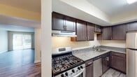 This single-floor residence offers a thoughtfully designed layout with a blend of practical finishes and comfortable living spaces. The main living area features durable gray wood-look flooring and warm neutral-toned walls, with a sliding glass door that fills the room with natural light and provides direct access to the outdoors. An accent wall in a soft blue-gray hue adds a touch of character to the living space. The open layout flows seamlessly into a secondary living or dining area, distinguished by lighter wood-tone flooring and a ceiling fan overhead, with a set of bi-fold closet doors adding convenient built-in storage. The kitchen is well-equipped with rich dark-espresso cabinetry, granite-look countertops, and a full suite of stainless steel appliances including a gas range, dishwasher, and refrigerator, complemented by a range hood and undermount sink. The bedroom features plush wall-to-wall carpeting and a large window with vertical blinds, allowing for natural light and privacy. The bathroom is clean and functional, offering a tub-shower combination with built-in shelving, wood-look flooring, and a vanity with granite-style countertops, maintaining a cohesive aesthetic throughout the home.