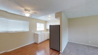 This single-floor space offers a functional and straightforward layout, with an open connection between the living area and kitchen that promotes a sense of flow throughout. The kitchen is outfitted with white cabinetry along the lower and upper sections, providing ample storage. A stainless-steel refrigerator anchors the kitchen visually, while a sink sits beneath a window that invites natural light into the space. The countertops are clean and practical, suited for everyday use. A partial wall separates the kitchen from the adjacent living area, adding subtle definition without fully closing off the space. The living area features hardwood-style flooring that transitions to carpet, and a ceiling fan overhead provides both lighting and air circulation. Neutral beige walls run throughout, creating a consistent and understated palette. Textured ceilings are present in both areas. Window blinds offer privacy while still allowing light to filter in. Overall, the space presents a modest, well-maintained interior with a no-frills, utilitarian character suited for comfortable day-to-day living.