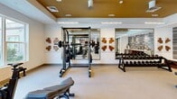 This single-floor fitness center in Richmond, Virginia, is a thoughtfully designed space that blends functionality with upscale aesthetics. The layout is organized to accommodate a comprehensive workout experience, with distinct zones dedicated to strength training, cardio, and recovery.

The strength training area is anchored by a power rack with a barbell, an adjustable bench, and a full dumbbell rack spanning a wide range of weights. Large mirrors line the walls, visually expanding the space and allowing for form monitoring during exercise. Framed wooden art pieces add a refined touch to the white walls, while a warm gold ceiling accent introduces an elevated design element.

The cardio zone features modern equipment including treadmills, ellipticals, and a rowing machine, all positioned near generous windows that invite natural light and offer views of the surrounding greenery — an inviting backdrop during workouts.

A standout feature is a dedicated refreshment station with warm wood cabinetry, a beverage refrigerator, open shelving, and a textured tile accent wall in neutral tones, adding a boutique feel to the space.

Glass-paneled doors lead to an adjacent room, adding to the sense of openness. Linear LED lighting throughout ensures bright, even illumination across the entire facility.