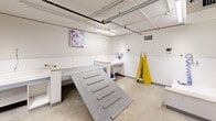 Located at 1225 Santa Fe Dr in Denver, CO, this purpose-built dog wash facility offers a compact, well-organized single-room space designed to accommodate all aspects of pet grooming. The room features durable concrete flooring and white-painted walls with a wainscoting-style lower panel, both chosen for easy cleaning and maintenance. Overhead, exposed utility piping, conduit, and fluorescent tube lighting run across the ceiling, giving the space a functional, no-frills industrial character. A standout feature is the textured walk-up ramp, which provides dogs of all sizes safe and easy access to the elevated grooming area. Built-in white cabinetry and countertops along the walls offer ample workspace and storage for grooming supplies, complemented by a dedicated wash station with water hookups and a flexible hose. Yellow grooming aprons hang conveniently on wall hooks, keeping essential equipment within easy reach. Two sturdy black doors provide entry and exit access, with visible fire suppression infrastructure and HVAC vents ensuring safety and comfort. Playful dog-themed artwork adorns the walls, adding a welcoming touch to an otherwise utilitarian environment. This facility is well-equipped to serve pet owners seeking a convenient, self-service grooming solution in the heart of Denver.