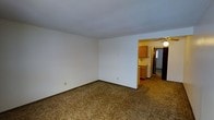 This single-floor residential space offers a clean, functional layout designed for comfortable everyday living. The main living area is finished with wall-to-wall carpeting in a warm, neutral tone, providing a cozy foundation throughout the space. Crisp white walls and a textured ceiling contribute to a bright, open atmosphere that feels airy and welcoming.

Visible from the living area, the kitchen connects seamlessly through an open doorway, creating a practical flow between the two spaces. The kitchen features classic honey-toned wood cabinetry that provides ample storage, and is outfitted with a ceiling fan for added ventilation. The cabinetry's warm finish adds a touch of traditional character to the overall aesthetic.

The open doorway framing between the living room and kitchen serves as a natural architectural divider, defining each area while maintaining an open, connected feel. The consistent carpeting and neutral color palette throughout create a cohesive, unified look that offers a versatile backdrop for a variety of interior styles. A hallway visible beyond the kitchen suggests additional rooms, further extending the functional footprint of this straightforward and practical single-floor layout.