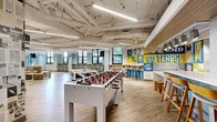 Located at 8204 Baltimore Ave in College Park, Maryland, this expansive single-floor community lounge offers a generously proportioned open-plan layout designed for recreation, socializing, and casual gathering. The space is defined by its energetic, university-spirited aesthetic, featuring a bold "Go Terps!" mural rendered in the University of Maryland's signature blue and gold that serves as a dramatic focal point throughout the main game area.

The floor plan flows seamlessly across several distinct activity zones. A dedicated game area houses foosball tables and a pool/ping-pong table, all set atop wood-look luxury vinyl flooring that runs throughout the entire space. A window-lined bar ledge with industrial-style stools runs along one wall, bathing the area in natural light and offering views of the surrounding neighborhood.

A standout architectural feature is a freestanding green wall enclosure — a lush, floor-to-ceiling living moss and foliage installation with a mounted flat-screen TV, creating a cozy lounge pod surrounded by plush modular seating and side tables. Nearby, a vertical open shelving unit with warm wood shelves adds a functional and decorative element.

Additional seating zones include upholstered modular benches and sofas, while nature-themed wall murals depicting bamboo and forest scenery add depth and a calming contrast to the vibrant game areas. Exposed ductwork and linear LED lighting on white ceilings contribute to the space's modern, urban feel.