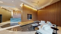 The lobby at 360 Huguenot Street in New Rochelle, NY presents a refined, contemporary design that makes a strong first impression. The entrance is defined by a sweeping curved glass curtain wall with dark metal framing, flooding the space with natural light and offering clear sightlines to the street beyond. Polished large-format marble tiles in contrasting tones of cream and deep charcoal define distinct zones across the floor, guiding movement through the space with understated elegance.

Warm, vertically grained wood paneling envelops the walls, creating a rich, welcoming ambiance that balances the cool tones of the stone and glass. A central freestanding wood partition houses a sleek, recessed fireplace with a low floating hearth, serving as a dramatic focal point that divides the entry corridor from the lounge area. The concierge station features a sculptural blue-veined marble desk set against the wood-paneled backdrop, accented by a delicate globe chandelier above.

The lounge area is furnished with low-profile white upholstered seating and minimalist side tables, positioned beneath a spectacular branching LED chandelier that evokes a constellation of light. Linear LED cove lighting traces the ceiling perimeter, adding a layer of ambient warmth throughout the lobby.