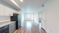 This single-floor unit features a clean, contemporary design characterized by white walls, wood-style laminate flooring, and an open-concept layout that connects the kitchen and living area seamlessly. The kitchen is efficiently designed with a two-tone cabinet scheme pairing white upper cabinets with warm wood-toned lower cabinets, complemented by granite-style countertops and a double stainless steel sink. Black appliances, including a full-size refrigerator, dishwasher, and range, complete the functional kitchen space.

The open living area extends naturally from the kitchen, offering a bright, flexible space illuminated by recessed lighting and natural light from windows fitted with horizontal blinds. The bedroom spaces maintain the same consistent flooring and neutral white walls, each benefiting from natural light through window openings.

The bathroom includes a vanity with granite-style countertops and a wood-toned cabinet base matching the kitchen's design language, accompanied by a large mirror, a grab bar, and wood-style flooring. A doorway from the bathroom leads to an adjacent walk-in closet, adding practical storage. Overall, the unit presents a cohesive, modern aesthetic with consistent finishes throughout.