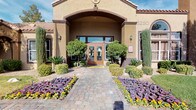 This single-story space makes a strong first impression with its well-maintained exterior, where a covered entryway framed by warm stucco walls and decorative tile roofing leads visitors along a paver walkway flanked by vibrant flowering plants, mature shrubs, and desert-adapted landscaping. Wall-mounted lanterns add an inviting, classic touch to the facade.

Inside, the space is anchored by a dramatic vaulted ceiling that amplifies the sense of openness and volume. A decorative chandelier serves as a focal point overhead, while recessed lighting ensures the interior remains bright and welcoming throughout. A large framed mirror mounted high on the accent wall further enhances the sense of depth and space.

The main lounge area is thoughtfully arranged with a pair of patterned wingback chairs, a plush sofa with coordinating accent pillows, and a round coffee table centered on a geometric area rug. The muted gray and blue color palette lends a contemporary yet comfortable feel. French doors along the back wall offer views of an outdoor pool area, filling the interior with natural light. A service counter alcove to one side suggests a functional reception or administrative area within the space.