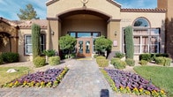 This single-story space makes a strong first impression with its well-maintained exterior, where a covered entryway framed by warm stucco walls and decorative tile roofing leads visitors along a paver walkway flanked by vibrant flowering plants, mature shrubs, and desert-adapted landscaping. Wall-mounted lanterns add an inviting, classic touch to the facade.

Inside, the space is anchored by a dramatic vaulted ceiling that amplifies the sense of openness and volume. A decorative chandelier serves as a focal point overhead, while recessed lighting ensures the interior remains bright and welcoming throughout. A large framed mirror mounted high on the accent wall further enhances the sense of depth and space.

The main lounge area is thoughtfully arranged with a pair of patterned wingback chairs, a plush sofa with coordinating accent pillows, and a round coffee table centered on a geometric area rug. The muted gray and blue color palette lends a contemporary yet comfortable feel. French doors along the back wall offer views of an outdoor pool area, filling the interior with natural light. A service counter alcove to one side suggests a functional reception or administrative area within the space.
