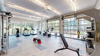 Located in Ocoee, FL, this fitness center is a modern, thoughtfully designed gym that offers a comprehensive workout environment. The space is defined by its bright, open layout, enhanced by large floor-to-ceiling glass garage doors that open toward an outdoor courtyard, inviting abundant natural light and views of lush Florida palm trees and landscaping. Additional full-panel windows along the perimeter reinforce the airy, energizing atmosphere.

The flooring features a light wood-style tile accented with blue geometric inlays, giving the space a clean, contemporary aesthetic. Overhead, linear fluorescent lighting provides consistent, even illumination throughout.

The cardio zone is well-appointed with multiple treadmills and elliptical machines, each equipped with individual digital screens, all positioned to take advantage of the scenic outdoor views. The strength training area includes stationary bikes, a cable/functional trainer machine, a weight bench, a full dumbbell rack, medicine balls, and a Reebok step platform. TRX suspension straps add versatile training options.

A bold branded mural anchors one wall, lending an motivational, gym-specific character to the space. A water fountain station is also conveniently accessible. Overall, this fitness center delivers a fully equipped, light-filled training environment suited for a wide variety of workout routines.