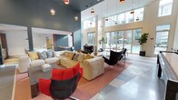 This contemporary community lounge is designed to impress from the moment you enter. The space is anchored by dramatic double-height ceilings that create an airy, open atmosphere, while a series of pendant lights with amber glass shades cascade from above, adding warmth and a touch of sophistication to the modern aesthetic. The walls are finished in a rich, deep gray-blue, providing a striking contrast to the bright, light-filled interior. Large floor-to-ceiling windows and glass doors line the far wall, flooding the space with natural light and offering seamless views of the outdoor pool and courtyard beyond. The lounge is thoughtfully furnished with a generous sectional sofa in neutral tones, complemented by bold red accent chairs and sleek black contemporary seating, creating a dynamic and inviting social environment. A tiled floor in a soft stone palette grounds the space and extends toward the pool-facing entrance. Greenery is subtly incorporated through potted plants, adding a natural, refreshing element. Whether used for casual gatherings or quiet relaxation, this lounge offers a polished, resort-inspired setting that balances comfort with modern design.