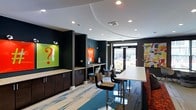 Located at 4451 Telfair Blvd in Camp Springs, Maryland, this single-floor commercial space makes a strong first impression with its bold, contemporary design. The interior is defined by a striking color palette of deep teal and dark charcoal walls, contrasted by crisp white ceilings that feature a curved, layered architectural detail with recessed lighting throughout. The flooring combines striped carpet and hard surface materials, adding visual texture and delineating different functional zones within the open layout. A built-in counter area with dark cabinetry and light granite-style surfaces provides a practical workstation or service zone, complemented by a vibrant green glass backsplash panel featuring graphic artwork. High bar-height communal tables with mixed seating, including patterned bar stools, create a casual yet functional gathering area. A secondary workstation with built-in shelving offers additional utility space. Large glass entry doors flood the interior with natural light and provide views to an exterior area beyond. Colorful artwork and branded signage contribute to the space's energetic and modern ambiance, making it well-suited for a hospitality lobby, co-working lounge, or customer-facing business environment.