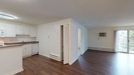 Unit 303 is a well-appointed 1-bedroom, 1-bathroom residence spread across a single floor. The open-concept living area is a defining feature of the unit, showcasing rich, dark hardwood-style flooring that extends throughout the main living spaces. Painted white walls and neutral tones create a clean, bright atmosphere, while a painted brick accent wall in the living area adds a subtle architectural character. A baseboard heating system and wall-mounted AC unit ensure year-round comfort. A louvered closet door provides convenient built-in storage within the main living space. The kitchen is efficiently designed with ample white cabinetry, a tile backsplash, and laminate countertops. White appliances include a refrigerator, electric range, built-in microwave, and dishwasher, making the space fully functional. A ceiling-mounted light fixture provides focused illumination. The bedroom, accessible from the main living area, features carpeted flooring and a ceiling fan for added comfort. The bathroom is finished with white tile, offering a full tub-shower combination with chrome fixtures, a vanity with mirror, and tile flooring, maintaining a clean and cohesive aesthetic throughout.