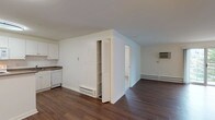 Unit 303 is a well-appointed 1-bedroom, 1-bathroom residence spread across a single floor. The open-concept living area is a defining feature of the unit, showcasing rich, dark hardwood-style flooring that extends throughout the main living spaces. Painted white walls and neutral tones create a clean, bright atmosphere, while a painted brick accent wall in the living area adds a subtle architectural character. A baseboard heating system and wall-mounted AC unit ensure year-round comfort. A louvered closet door provides convenient built-in storage within the main living space. The kitchen is efficiently designed with ample white cabinetry, a tile backsplash, and laminate countertops. White appliances include a refrigerator, electric range, built-in microwave, and dishwasher, making the space fully functional. A ceiling-mounted light fixture provides focused illumination. The bedroom, accessible from the main living area, features carpeted flooring and a ceiling fan for added comfort. The bathroom is finished with white tile, offering a full tub-shower combination with chrome fixtures, a vanity with mirror, and tile flooring, maintaining a clean and cohesive aesthetic throughout.