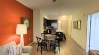 This single-floor residence showcases an open-concept design that integrates the living, dining, and kitchen areas into one cohesive space. A striking burnt-orange accent wall anchors the living area, adding warmth and visual interest against the predominantly white walls. Light wood-tone flooring runs throughout, contributing to a bright, airy atmosphere.

The dining area features a round glass-top table with black cross-back chairs, positioned centrally to take advantage of the open layout. A ceiling fan overhead provides both function and subtle style. Adjacent to the dining space, the kitchen is outfitted with dark cabinetry, stainless steel appliances including a French door refrigerator, and a peninsula with a light-colored countertop and bar seating. A pendant light above the peninsula adds a modern touch.

A hallway extends toward the entrance, lined with white doors and framed artwork, giving the space a clean, organized feel. Track lighting and recessed fixtures ensure the space is well-lit throughout. Overall, the unit offers a modern, functional layout with thoughtful design details that balance comfort and contemporary style.