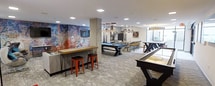 This single-floor entertainment space is thoughtfully designed to deliver a vibrant social experience through a blend of industrial and modern aesthetics. A striking exposed brick accent wall, adorned with colorful mural artwork, serves as a dramatic focal point and anchors a bar-height counter complete with a wooden top and red metal barstools — perfect for casual gathering and conversation. A wall-mounted flat-screen television completes the lounge vibe in this area. Moving deeper into the space, a full-sized pool table takes center stage beneath an eye-catching arrangement of decorative pendant chandeliers, adding both elegance and character to the room. A shuffleboard table and additional game surfaces invite friendly competition and extended play. A high-top bar counter with yellow barstools defines a secondary social zone, providing additional seating and versatility. Large windows along the perimeter flood the space with natural light, enhancing the open, airy feel of the room. Recessed ceiling lighting complements the ambient glow throughout. The flooring features a patterned carpet that adds warmth while tying together the eclectic design elements. Overall, the space strikes a confident balance between energetic entertainment and refined style.