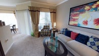 This single-floor living space showcases a thoughtfully designed interior that balances comfort and style. The living area is highlighted by elegant crown molding that frames the ceiling, adding a refined architectural detail to the room. Large windows dressed with layered gold and brown drapery allow generous natural light to fill the space, creating a warm and inviting ambiance. A sliding glass door provides access to an outdoor area, further enhancing the flow of natural light.

The room features plush wall-to-wall carpeting that adds a soft, comfortable feel underfoot. Seating is arranged around a sleek oval glass-top coffee table with a dark wood frame, which serves as a focal point in the living area. A neutral-toned sofa is accented with bold blue and red throw pillows, while a patterned accent chair in blue and yellow adds a lively contrast. A bold abstract artwork in red and yellow tones anchors the wall above the sofa, adding a dynamic visual element. Decorative accessories, including teal glass candleholders and a woven storage basket, complete the polished, curated look of the space.