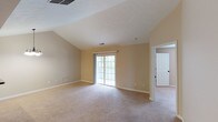 This single-floor space offers a bright and open layout defined by its notable vaulted ceiling, which adds a sense of height and airiness to the interior. The main living area is finished with soft, neutral-toned wall-to-wall carpeting that contributes to a warm and comfortable ambiance throughout. A large sliding glass door serves as a central architectural feature, inviting an abundance of natural light into the space while providing direct views of the surrounding greenery outdoors. The walls are painted in a gentle beige tone, complementing the light-colored carpet and creating a cohesive, understated aesthetic. A dedicated HVAC vent is integrated into the ceiling, ensuring climate control throughout the room. White painted trim and baseboards frame the space cleanly, adding a crisp contrast to the warm wall tones. An adjacent doorway with a paneled white door leads to a secondary room, suggesting a functional and well-connected floor plan. The textured ceiling adds a subtle design detail overhead. Overall, the space presents a clean, move-in-ready environment with classic residential finishes and a layout that emphasizes openness and natural illumination.