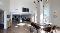 This single-floor space showcases a contemporary open-plan design characterized by dramatically high ceilings and crisp white walls that amplify the sense of volume and airiness throughout. Large windows along the perimeter flood the interior with natural light, casting warm sunlight across the wide-plank hardwood flooring. The layout is thoughtfully organized into distinct functional zones while maintaining a cohesive, flowing atmosphere. A bar-height counter with sleek metal surfaces and brown leather-upholstered stools provides a casual seating area ideal for informal meetings or individual work. Pendant lighting fixtures with an industrial aesthetic hang overhead, adding both task illumination and visual interest. Further into the space, lounge-style seating with a yellow sofa and accent chairs creates a relaxed gathering area. A wall-mounted flat-screen TV serves as a central media feature. Toward the rear, additional seating arrangements and what appears to be a kitchenette or service area offer further functionality. Decorative wall art and sculptural accents contribute personality to the otherwise minimalist, modern aesthetic throughout the space.