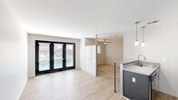 Located in Royal Oak, MI 48067, this efficiently designed single-floor unit offers a bright and modern living experience. The open-concept layout connects the living area and kitchen seamlessly, maximizing the use of available space. Light wood-tone flooring runs throughout the main living area, contributing to a clean, contemporary aesthetic.

The living space is anchored by a striking set of black-framed glass French doors that invite abundant natural light and provide outdoor views, creating an airy atmosphere. A semi-transparent partition wall subtly defines the adjacent carpeted room, which features a ceiling fan for added comfort.

The kitchen is thoughtfully appointed with a granite-topped peninsula that doubles as a breakfast bar, complemented by pendant lighting in brushed nickel. Dark cabinetry on the peninsula contrasts with white upper cabinets above the cooking area. Black appliances — including a refrigerator, electric range, and dishwasher — complete the functional kitchen suite, while a stainless steel sink adds a polished touch.

The bathroom features floor-to-ceiling white subway tile in the shower enclosure, delivering a clean and timeless finish. Throughout the unit, white walls and neutral tones create a cohesive, understated palette well-suited for modern living in the heart of Royal Oak.