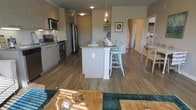 This single-floor space showcases a thoughtfully designed open-concept layout that seamlessly connects the kitchen, dining, and living areas. The kitchen is well-appointed with stainless steel appliances, including a range, microwave, and refrigerator, all complemented by sleek cabinetry in a neutral gray-beige palette. A central kitchen island serves as the focal point, offering bar-style seating with light teal chairs and pendant lighting above, adding a warm and inviting ambiance. Built-in shelving on the adjacent wall provides both functional storage and display space. Warm hardwood-style flooring flows throughout, tying the open areas together cohesively and lending a clean, contemporary feel. The living area extends naturally from the kitchen and dining space, with natural light filtering in to brighten the interior. Soft, neutral wall tones enhance the sense of openness, while decorative accents and colorful textiles introduce personality and warmth. The layout is efficient and practical, making excellent use of the available space while maintaining an airy, comfortable atmosphere. A doorway visible in the background hints at additional rooms, completing this well-organized and modern single-floor residence.