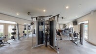 This fitness center is a spacious, single-floor facility designed to support a wide range of workout routines. The layout is thoughtfully organized, with distinct zones for cardiovascular training and strength conditioning. Along one side of the room, a row of Cybex cardio machines — including treadmills and stationary bikes — lines the wall beneath mounted flat-screen televisions, creating an engaging environment for endurance training. Large windows and glass doors along this stretch admit generous natural light while offering views of the surrounding outdoor area.

The strength-training section features a robust collection of equipment, including cable machines, a functional trainer, selectorized weight machines, and a well-stocked dumbbell rack. A dedicated free-weight area with benches and a squat rack provides options for resistance training. Full-length mirrors are strategically placed throughout, enhancing the sense of openness while allowing users to monitor their form.

Recessed lighting and ceiling fans maintain a comfortable atmosphere throughout. The flooring consists of durable, cushioned tile throughout the space. The neutral color palette of warm whites and grays lends the environment a clean, professional aesthetic. Overall, the fitness center is designed to accommodate a broad range of fitness needs in a well-lit, organized, and functional setting.