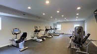 This fitness center is thoughtfully designed to support a comprehensive cardio-focused workout experience. The space is outfitted with a lineup of True-brand exercise equipment, including elliptical trainers, treadmills, a recumbent bike, and a cable resistance machine, all arranged to allow comfortable movement between stations. The rubber flooring provides both durability and safety underfoot, making it well-suited for high-traffic athletic use. Recessed ceiling lights are evenly distributed throughout, ensuring consistent, glare-free illumination across the entire floor. The neutral gray walls contribute to a clean, modern aesthetic that keeps the focus on functionality. Natural light filters in through windows positioned along one wall, adding a welcoming brightness to the environment. The open layout provides ample space between machines, promoting ease of use and a comfortable workout atmosphere. Overall, the fitness center presents a practical and well-maintained setting suitable for daily exercise routines.