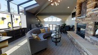 This single-floor space makes a striking architectural statement with its soaring vaulted ceilings that draw the eye upward, creating an expansive and airy atmosphere. Multiple skylights are positioned along the roofline, flooding the interior with abundant natural light throughout the day and enhancing the sense of openness.

The living area serves as the centerpiece of the space, anchored by a curved, tufted gray sectional sofa accented with mustard yellow and neutral throw pillows. A round glass-top coffee table with a sculptural metallic base sits at the heart of the seating arrangement, adding a contemporary touch. A stone-clad feature wall adds warmth and texture, providing a refined architectural contrast to the sleek surroundings.

The open layout transitions seamlessly into a dining area, where a dark-toned table and upholstered chairs sit beneath the continued vaulted ceiling. A sputnik-style chandelier introduces a mid-century modern element, complementing the wall sconces that provide ambient warmth. A set of glass doors at the far end allows natural light to pour in while offering a visual connection to the outdoors, completing the cohesive and sophisticated design of the space.