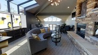 This single-floor space makes a striking architectural statement with its soaring vaulted ceilings that draw the eye upward, creating an expansive and airy atmosphere. Multiple skylights are positioned along the roofline, flooding the interior with abundant natural light throughout the day and enhancing the sense of openness.

The living area serves as the centerpiece of the space, anchored by a curved, tufted gray sectional sofa accented with mustard yellow and neutral throw pillows. A round glass-top coffee table with a sculptural metallic base sits at the heart of the seating arrangement, adding a contemporary touch. A stone-clad feature wall adds warmth and texture, providing a refined architectural contrast to the sleek surroundings.

The open layout transitions seamlessly into a dining area, where a dark-toned table and upholstered chairs sit beneath the continued vaulted ceiling. A sputnik-style chandelier introduces a mid-century modern element, complementing the wall sconces that provide ambient warmth. A set of glass doors at the far end allows natural light to pour in while offering a visual connection to the outdoors, completing the cohesive and sophisticated design of the space.
