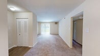 Located at Wellington Lakes in Fredericksburg, Virginia, this one-bedroom, one-bathroom residence offers a practical and well-organized single-floor layout. The combined dining and living area is the heart of the home, featuring neutral-toned carpeting, bright white walls, and a large sliding glass door dressed with vertical blinds that fills the space with natural light and provides an outdoor view. A pass-through window connects the living area directly to the kitchen, enhancing the open feel between spaces. The kitchen is outfitted with white cabinetry, light-toned countertops, wood-style vinyl flooring, and a full suite of white appliances including a gas range, dishwasher, and refrigerator. An adjacent pantry provides additional storage. A hallway leads from the main living area to the bedroom, which is generously sized and carpeted, with a window allowing in natural light and two closets — including a louvered bifold closet and a separate walk-in style closet — offering ample storage. The bathroom features a clean, all-white tub-shower combination with built-in shelving. Throughout the residence, the neutral color palette and functional layout create a clean, move-in-ready atmosphere.
