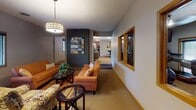 Located in Coon Rapids, MN, this single-floor community amenity space offers a thoughtfully designed environment for relaxation, socializing, and fitness. The lounge area serves as the heart of the space, anchored by warm orange upholstered seating arranged around a glass-top coffee table atop a boldly striped area rug. A dark-toned decorative brick accent wall provides striking contrast against the neutral gray walls, while a geometric drum pendant light and a framed mixed-media artwork add character and visual interest. Interior windows with wood trim frame views into the adjacent fitness center, creating a sense of openness and connectivity throughout. The dining area features multiple high-top tables with bar-height stools beneath a matching pendant fixture, making it well-suited for casual gatherings. Built-in wooden cabinetry lines one wall, offering ample storage, and a side door provides convenient access. The fitness center, visible through the interior windows, is equipped with cardio machines including a treadmill and stationary bike, along with free weights and exercise mats, all set within a well-lit, carpeted room with natural light from exterior windows. Textured ceilings and warm wood trim details carry throughout, giving the overall space a cohesive and comfortable feel.