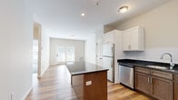 This thoughtfully laid-out one-bedroom, one-bathroom residence offers a comfortable and functional living environment across a single floor. The open-concept kitchen and living area forms the heart of the home, where warm hardwood-style flooring flows seamlessly throughout. The kitchen is well-appointed with stainless steel appliances, including a refrigerator, dishwasher, range, and microwave. A central island with a dark granite countertop provides additional prep and dining space, while a two-tone cabinetry scheme — white upper cabinets paired with rich wood-toned lower cabinets — adds visual depth. A white subway tile backsplash completes the polished look. The living area is bright and inviting, benefiting from natural light that streams in through French doors. The bedroom offers a cozy retreat with plush carpeting, a large window with views to the outdoors, and clean white panel doors. The bathroom features a spacious walk-in shower with sliding glass doors, built-in shelving, chrome fixtures, and granite vanity countertops, all set atop neutral tile flooring. Hallways connect the rooms with ease, contributing to a practical and cohesive floor plan throughout the residence.
