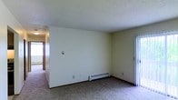 This single-floor space presents a straightforward and functional layout characterized by clean, neutral white walls and wall-to-wall carpeting throughout. The main living area is generously proportioned, providing ample room for flexible furniture arrangements. A textured ceiling adds a subtle design element, while the open corner configuration of the room creates a sense of spaciousness.

A baseboard heater runs along the base of the wall, serving as the primary heat source for the space. A light switch panel is centrally positioned on one wall, contributing to the room's practical design. To the right, a sliding door with vertical blinds offers access to an adjacent area or exterior space, allowing for natural light to filter into the room.

A hallway visible toward the left side of the space connects to additional rooms or areas, hinting at a functional floor plan. The overall ambiance is understated and neutral, providing a blank canvas that can be adapted to a variety of uses and personal styles. The space is well-suited for those seeking a practical, move-in-ready environment with essential amenities already in place.