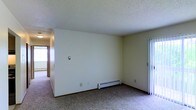 This single-floor space presents a straightforward and functional layout characterized by clean, neutral white walls and wall-to-wall carpeting throughout. The main living area is generously proportioned, providing ample room for flexible furniture arrangements. A textured ceiling adds a subtle design element, while the open corner configuration of the room creates a sense of spaciousness.

A baseboard heater runs along the base of the wall, serving as the primary heat source for the space. A light switch panel is centrally positioned on one wall, contributing to the room's practical design. To the right, a sliding door with vertical blinds offers access to an adjacent area or exterior space, allowing for natural light to filter into the room.

A hallway visible toward the left side of the space connects to additional rooms or areas, hinting at a functional floor plan. The overall ambiance is understated and neutral, providing a blank canvas that can be adapted to a variety of uses and personal styles. The space is well-suited for those seeking a practical, move-in-ready environment with essential amenities already in place.