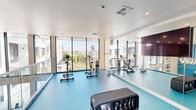 Located at 525 N 7th St in San Jose, California, this modern fitness center offers a thoughtfully designed exercise space that combines functionality with contemporary style. The room is defined by striking turquoise interlocking floor tiles that run throughout, providing both a vibrant aesthetic and a durable, cushioned workout surface.

Floor-to-ceiling glass windows line one side of the room, flooding the space with abundant natural light and offering open views of the surrounding San Jose cityscape, complete with tree canopies and neighboring rooftops. A sleek glass railing system runs along the open interior edge, adding to the modern, airy feel of the space.

The exercise room is well-equipped with cardio machines including stationary cycling bikes and elliptical trainers, along with TRX suspension training systems and free weights. A large wall-mounted mirror reflects the room and enhances the sense of openness, while a mounted flat-screen display provides entertainment or instructional content during workouts.

Recessed ceiling lighting supplements the generous natural light, ensuring the space is well-lit at all hours. A contemporary decorative light fixture serves as a design focal point near the glass railing. Clean white walls and a crisp white ceiling complete the polished, minimalist aesthetic, making this fitness center an energizing and inviting environment for exercise.