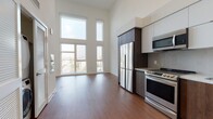 This two-story loft-style space showcases a striking contemporary design defined by its dramatic double-height ceilings and an expansive wall of floor-to-ceiling windows that flood the main living area with abundant natural light and offer open urban views. The rich, dark hardwood flooring throughout the main level adds warmth and contrast to the crisp white walls, creating a balanced modern aesthetic.

The open-plan kitchen is well-appointed with a full suite of stainless steel appliances, including a French door refrigerator, electric range, built-in microwave, and dishwasher. A combination of white upper cabinetry and dark wood lower cabinets is complemented by a herringbone-patterned tile backsplash, lending texture and visual interest to the space.

A staircase leads to the upper level, with a built-in shelving unit tucked neatly beneath it, maximizing storage. A glass door provides access to an outdoor area, further enhancing connectivity with the exterior. The bathroom features a dark wood vanity with a stone-topped countertop, an illuminated LED mirror, and light tile flooring, offering a clean and refined finish consistent with the overall design language of the space.