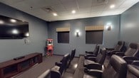 This dedicated theater space delivers a true cinematic experience within a well-appointed, single-level layout. The room is finished with deep blue-gray walls and a drop-tile ceiling with recessed lighting, creating an intimate, darkened ambiance ideal for screen viewing. Two large projection screens, each framed by striped valances, are mounted along the front wall, offering flexible viewing arrangements for audiences of varying sizes. Rows of plush, black leather recliner seats with built-in cup holders are arranged in a tiered, stadium-style configuration, ensuring clear sightlines from every position. Wall-mounted sconces provide soft accent lighting along the sides, complementing the overall moody atmosphere. A classic red-and-gold popcorn machine on a wheeled cart adds a charming, nostalgic touch reminiscent of a traditional movie house. Patterned low-pile carpeting covers the floor throughout, contributing to the room's acoustic comfort and refined aesthetic. A media console along one wall provides additional functionality. Altogether, this theater space is thoughtfully designed to offer an engaging, comfortable, and fully immersive entertainment environment.