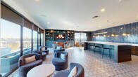 This thoughtfully designed single-floor amenity lounge offers a variety of distinct zones suited for socializing, relaxing, and entertaining. The main lounge area is generously proportioned, featuring patterned tile flooring and an arrangement of plush, curved barrel chairs in deep blue-gray tones grouped around low tables. A teal built-in shelving unit anchors one wall, framing a fireplace display and flat-screen TV, while large floor-to-ceiling sliding glass doors flood the space with natural light and provide direct access to an outdoor terrace with open skyline views.

The bar and kitchen area serves as a central hub, distinguished by a striking floor-to-ceiling herringbone blue tile accent wall illuminated by a row of warm globe pendant lights. A marble-topped island with dark wood cabinetry provides ample counter space, complemented by bar seating along its edge. Adjacent to the bar, a compact dining nook features a mosaic tile accent wall, hardwood flooring, and a high-top table with upholstered counter stools.

A dedicated media lounge offers a more intimate retreat, with warm wood-paneled walls, a bold orange sectional sofa, a patterned area rug, and a wall-mounted flat-screen TV. A billiards table is also visible within the space. Throughout, recessed lighting and carefully curated décor maintain a cohesive, upscale atmosphere.