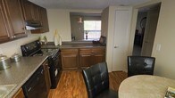 This two-floor residence offers a functional and cohesive layout that blends the kitchen, dining, and living areas into a connected living space. The kitchen is outfitted with dark wood cabinetry, a granite-style countertop, and black appliances, including an electric range. A peninsula counter serves as a natural divider between the kitchen and the adjacent living area, providing additional prep and serving space while maintaining an open feel. Warm hardwood flooring runs throughout, adding a rich, inviting tone to the main level. The dining area is positioned just off the kitchen, accommodating a dining set with tufted leather chairs. Beyond the peninsula, the living space benefits from a large window that invites natural light and provides a view of the outdoors, brightening the interior. Neutral wall tones complement the darker cabinetry and flooring, creating a balanced visual contrast throughout. A textured ceiling adds subtle architectural detail. The overall ambiance is warm and practical, with a layout designed to support everyday living across two floors.