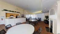 This single-floor gathering space is thoughtfully designed to accommodate a variety of events and social functions. The main area is generously sized, featuring rich brick-patterned tile flooring throughout and soaring vaulted ceilings accented by ceiling fans that enhance the open, airy atmosphere. Multiple round white tables with folding chairs are arranged throughout, providing flexible seating configurations for groups of varying sizes. A wall of floor-to-ceiling vertical blinds along one side floods the space with natural light while offering privacy as needed.

A dedicated lounge area anchors one end of the room, featuring an elegant fireplace framed by a decorative arch, complemented by artwork and tall potted plants that add warmth and greenery. A large flat-screen display is mounted nearby, making the space suitable for presentations or screenings.

The full kitchen is set behind a wide granite-topped white counter with decorative columns, equipped with wood cabinetry, a stainless steel refrigerator, a range, and a range hood. Greenery drapes over the kitchen partition, adding a charming decorative touch. A checkered tile entryway with French doors provides a distinct transition point. Additional folding tables along the windowed perimeter offer supplemental surface space for events.