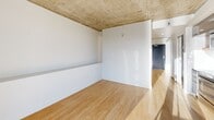Located at 22 NE 2nd Ave in Portland, Oregon, this efficiently designed unit offers a modern, urban living experience on a single floor. The space features warm bamboo hardwood flooring throughout, contrasted by striking exposed concrete ceilings that lend an industrial yet refined aesthetic. The main living area is anchored by dramatic floor-to-ceiling windows that frame breathtaking panoramic views of the Portland skyline, the Willamette River, and a major bridge corridor, flooding the interior with abundant natural light. A sleek, built-in kitchen is thoughtfully recessed into the wall, outfitted with glossy gray cabinetry, stainless steel appliances including a range and microwave, a chevron-patterned tile backsplash, and a warm wood open shelf that adds a natural accent. The bathroom is appointed with a stainless steel countertop vanity, a matte black undermount sink, a large mirror, and a natural wood shelf, combining industrial and organic materials seamlessly. A stacked washer and dryer unit provides convenient in-unit laundry. Clean white walls and a minimalist layout maximize the sense of openness throughout this well-appointed urban retreat.