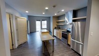 This single-floor space showcases an open-concept layout that seamlessly integrates a fully equipped kitchen with the surrounding living area. The kitchen is outfitted with stainless steel appliances, including a range with an electric cooktop, a large refrigerator, and a built-in microwave, all complemented by light wood cabinetry and open shelving above. A prominent stainless steel range hood serves as a functional focal point above the cooking area. A long, freestanding prep table with metal legs runs centrally through the space, offering ample workspace and a defining visual element. Dark hardwood flooring extends throughout, providing a warm contrast to the cool blue-gray walls. Recessed lighting illuminates the interior evenly, enhancing the clean, modern aesthetic. Toward the far end of the space, a set of windows allows natural light to filter in, brightening the open living area beyond the kitchen. Louvered doors on one side suggest additional storage or utility access. The overall ambiance is functional and contemporary, suited for both food preparation and social activity.