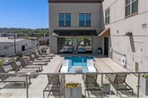 This rooftop spa and pool deck offers a well-appointed outdoor retreat set against an open urban skyline. The space centers on a rectangular pool with vivid turquoise water accented by mosaic tile detailing along its edges. Adjacent to the main pool, a dedicated hot tub provides a separate soaking area, while a pair of sleek white in-water lounge chairs allow guests to recline directly in the shallow water—a standout design feature that adds a resort-style sensibility to the space.

The surrounding deck is finished in a neutral concrete surround that keeps the aesthetic clean and modern. Generous rows of woven chaise lounges are arranged throughout the sunning area, offering ample seating for relaxation. Glass panel railings line the perimeter, preserving unobstructed sightlines across the surrounding cityscape and treetops under open sky.

The adjoining building facade is finished in light-toned paneling with large floor-to-ceiling glass garage-style doors, creating a seamless visual connection between the indoor amenity spaces and the outdoor deck. Outdoor showers and safety equipment are also present. The overall ambiance is bright, airy, and contemporary, well-suited for both active recreation and leisurely relaxation.