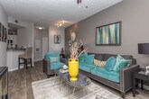 This single-floor residence offers a thoughtfully designed layout with a cohesive contemporary aesthetic carried throughout each space. The living room serves as the centerpiece of the home, featuring wood-style flooring, neutral gray walls, and a ceiling fan with integrated lighting. Natural light filters in through multiple windows dressed with layered dark curtains, creating a warm yet sophisticated atmosphere. A mix of teal, charcoal, and yellow accents adds a bold, curated feel to the space.

The bedroom continues the design palette with plush carpeting, a ceiling fan, and a generously sized window that allows natural light to fill the room. Decorative mirrors above the headboard wall add depth and visual interest, while dark drapery frames the window elegantly.

The kitchen is outfitted with white shaker-style cabinetry, dark countertops, and a double stainless steel sink, providing a clean, functional workspace with ample storage including a tall pantry-style cabinet.

The bathroom features a speckled gray countertop with an undermount sink, a large mirror, and a tiled shower stall, all finished in neutral tones that contribute to the space's calm and practical character.