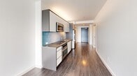 This thoughtfully designed one-bedroom, one-bathroom residence offers a clean, modern aesthetic across a single floor. The open-plan living and kitchen area is bright and airy, anchored by wide-plank hardwood-style flooring in a warm grey tone that flows seamlessly throughout the space. The contemporary kitchen runs along one wall and is equipped with stainless steel appliances — including a built-in oven, microwave, and gas cooktop — complemented by sleek grey cabinetry and a teal glass-tile backsplash that adds a refined pop of color. A large picture window at the end of the main living area frames sweeping views of the waterfront and city skyline, flooding the space with natural light. The bedroom is a calm, well-proportioned retreat, featuring floor-to-ceiling windows that continue the waterfront vistas and a set of sliding built-in closet doors with chrome detailing. The bathroom is finished with large-format stone-effect wall tiles surrounding a soaking tub and shower combination, accented by polished chrome fixtures and clean white surfaces. Crisp white walls throughout the residence create a bright, minimalist backdrop, making the space feel open, functional, and move-in ready.
