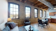 This single-floor loft showcases an impressive blend of industrial heritage and contemporary design. The open-concept living and dining area is the heart of the space, anchored by a striking wall of original exposed brick and a row of tall, arched windows with dark frames that flood the room with natural light and offer views of the surrounding neighborhood. Overhead, richly stained timber beams span the ceiling alongside exposed ductwork, reinforcing the industrial character throughout.

The living area features light hardwood flooring, while the bedroom transitions to plush carpet and continues the exposed brick aesthetic with similarly soaring wood-beam ceilings. The bedroom is generously proportioned, accommodating a large bed with ease, and includes a cozy seating nook and access to a private en-suite bath.

The kitchen is well-appointed with warm wood cabinetry, light quartz countertops, stainless steel appliances including a dishwasher and range, and a deep double sink — offering both style and functionality. The bathroom is clean and modern, finished with large-format tile flooring, a vanity matching the kitchen's cabinetry tones, and a shower-tub combination. A stacked in-unit washer and dryer adds everyday convenience.

Overall, this loft delivers a compelling combination of historic architectural detail and modern livability.