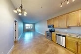Located at 721 N Clinton St in Syracuse, NY, this single-floor, one-bedroom, two-bathroom unit offers a well-organized layout with a comfortable sense of space. The open-concept kitchen and living area forms the heart of the unit, featuring polished concrete flooring that reflects natural light from the living room window. A decorative chandelier adds a warm focal point overhead. The kitchen is well-equipped with warm-toned maple cabinetry extending to the ceiling, granite-look countertops, and a full suite of stainless steel appliances including a range, over-the-range microwave, dishwasher, and refrigerator. Track lighting illuminates the kitchen workspace effectively. The spacious living area provides ample room for various furniture arrangements, with a wall-mounted heating unit beneath the front-facing window. The bedroom features soft carpeting, neutral-toned walls, and a window with sheer curtains that invite natural light and outdoor views of surrounding trees. Two closets provide practical storage solutions, one of which houses the tankless water heater and wire shelving. The unit includes two bathrooms, rounding out a functional and efficient floor plan suited for comfortable everyday living.