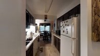 Located at 7 Elk Dr in Monticello, NY, this single-floor, two-bedroom, one-bathroom home offers a functional and cozy layout. The galley-style kitchen is a central feature, finished with dark cabinetry on both sides that provides ample storage. Light countertops contrast the dark cabinet tones, and the kitchen is equipped with a gas range, full-size refrigerator, and a sink positioned along the window-facing wall. The tile flooring runs throughout the kitchen, adding durability and easy maintenance underfoot. The kitchen opens directly into a dedicated dining area, creating a seamless flow between meal preparation and dining. The dining space is anchored by a table and chair set and is illuminated by a ceiling fan with an integrated light fixture. A window in the dining area invites natural light into the space and offers a view to the outside. The warm, lived-in ambiance of the home is complemented by decorative touches throughout. The single-floor layout ensures easy accessibility and efficient use of space, making this home a practical and comfortable residence in the Monticello community.