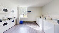 Located at 10 Highland Crescent in Kitchener, Ontario, this communal laundry facility offers a practical and welcoming environment designed to serve residents efficiently. The single-floor space is thoughtfully divided into distinct functional zones, all unified by light wood-style flooring, bright white walls, and a clean, modern aesthetic throughout.

The main laundry area is well-equipped with a full row of front-loading washing machines and dryers, including stacked units that make efficient use of vertical space. A utility sink provides added convenience, while multiple stainless steel folding tables offer ample surface area for sorting and organizing laundry. Bright fluorescent lighting ensures the workspace is well-illuminated at all times.

Natural light filters through a large window in the washing area, lending a pleasant, airy feel to what might otherwise be a purely utilitarian space. Waste and recycling bins are conveniently positioned throughout.

A separate waiting and common area provides a comfortable place to relax, furnished with modern seating, round tables, and chairs in a contemporary Scandinavian style. Ceiling-mounted light fixtures cast a warm glow over the space, which is further enlivened by decorative artwork and a potted plant. Additional seating near the entrance corridors and a payment or information station complete this thoughtfully designed, resident-focused facility.